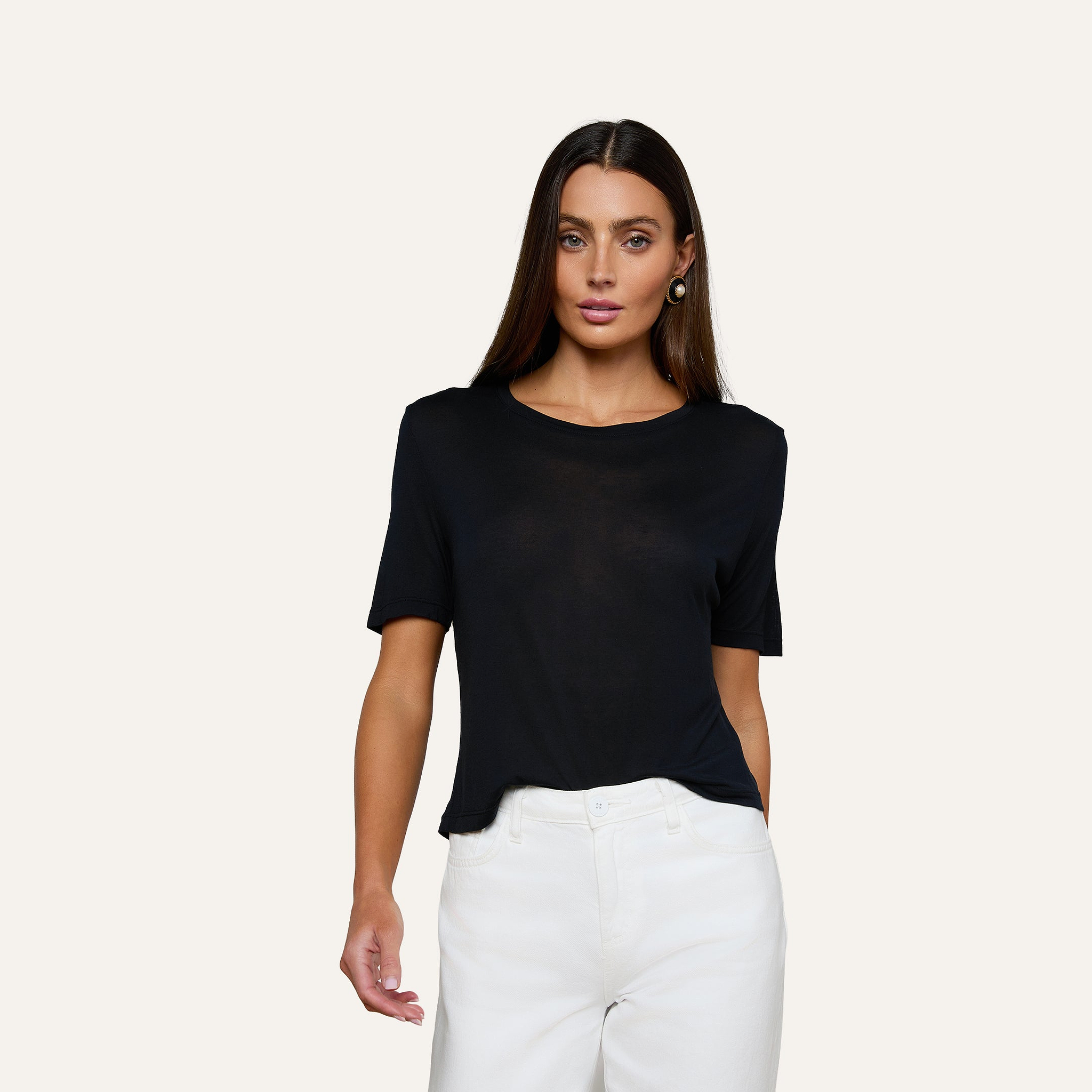 A woman with long brown hair wears the Remy Tee—a black, relaxed-fit off-duty top—paired with white pants, standing against a plain light-colored background.