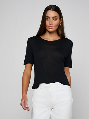 A woman with long brown hair wears the Remy Tee, a black relaxed fit tee made from lightweight jersey, paired with white pants. She stands against a plain light gray background, gazing forward with a neutral expression.