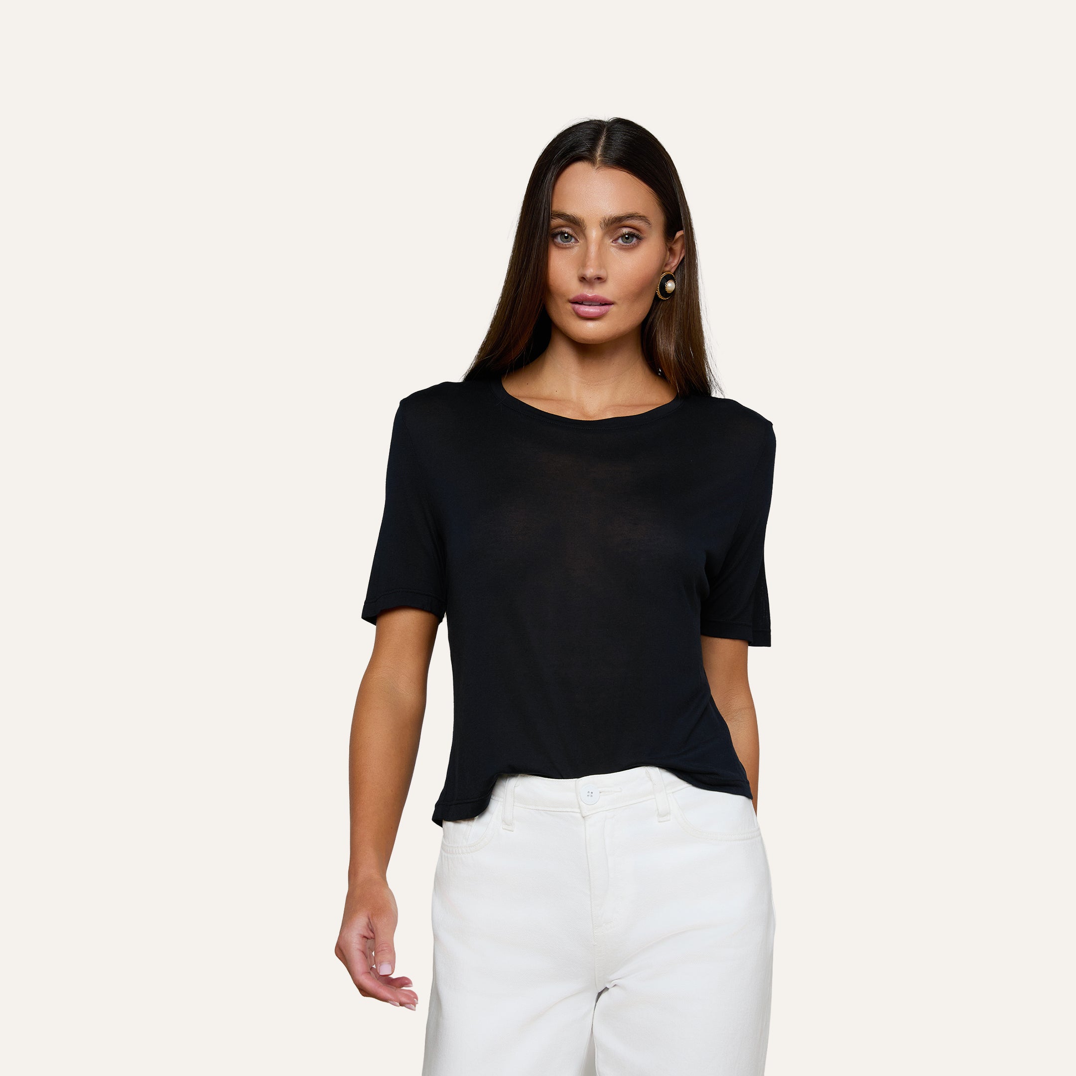 A woman with long brown hair wears the Remy Tee—a black, relaxed-fit off-duty top—paired with white pants, standing against a plain light-colored background.