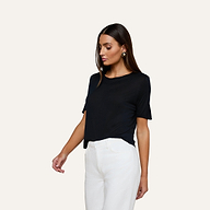 A woman stands against a plain background wearing the Remy Tee—a black, short-sleeve, relaxed fit top—paired with high-waisted white pants. She looks slightly downward, with one arm relaxed by her side and the other arm slightly bent.