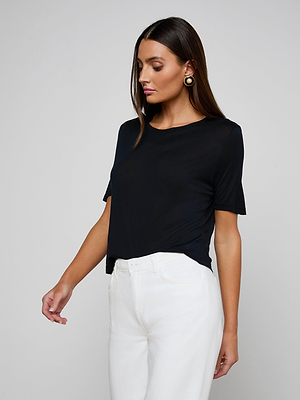 A woman with long brown hair wears the black Remy Tee in a relaxed fit with high-waisted white pants, standing against a plain light gray background and looking down to her left.