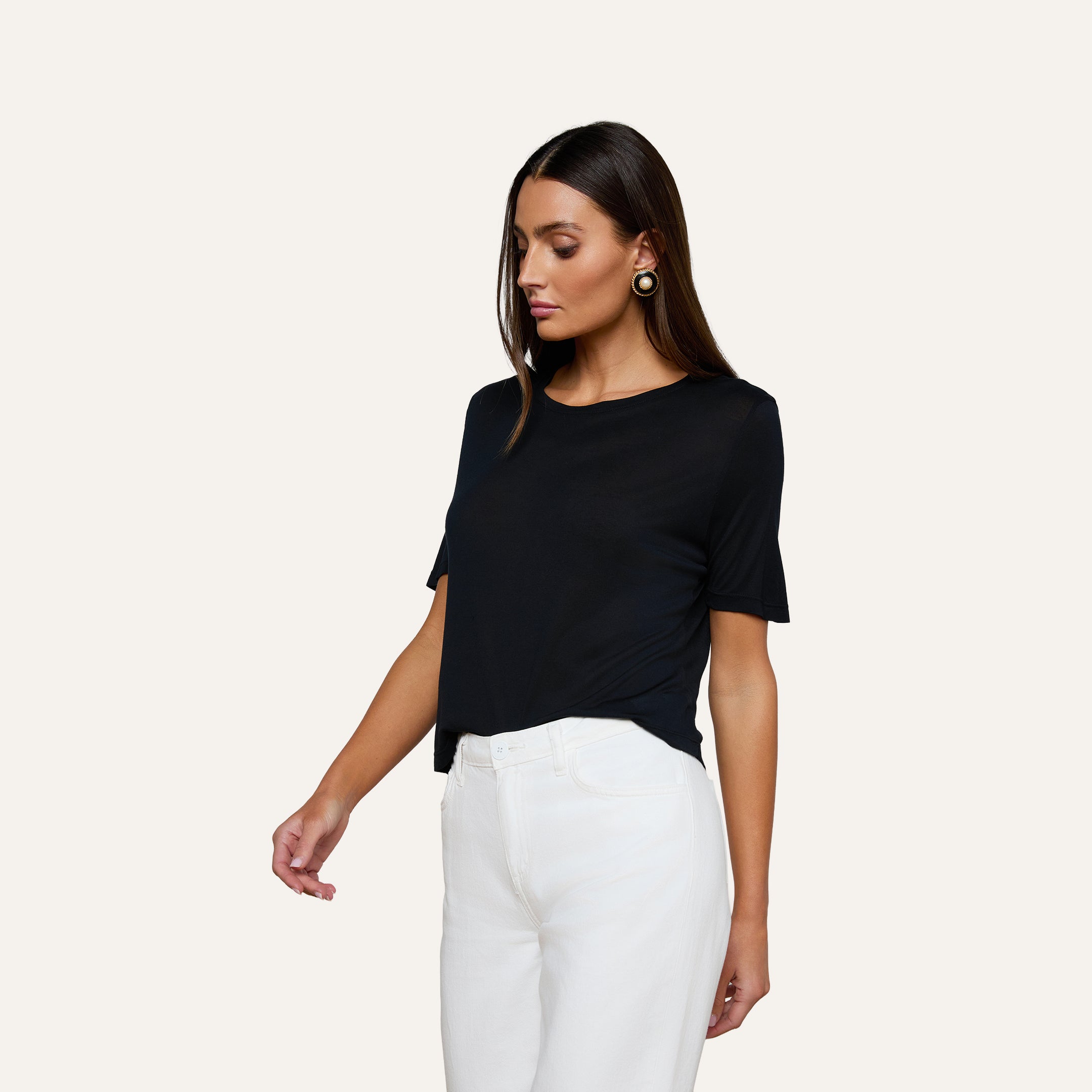 A woman stands against a plain background wearing the Remy Tee—a black, short-sleeve, relaxed fit top—paired with high-waisted white pants. She looks slightly downward, with one arm relaxed by her side and the other arm slightly bent.