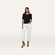 A woman stands against a plain white background, wearing the Remy Tee—a black, short-sleeve top with a relaxed fit—paired with white wide-leg pants and black high-heeled sandals. One hand is in her pocket as she looks directly at the camera.