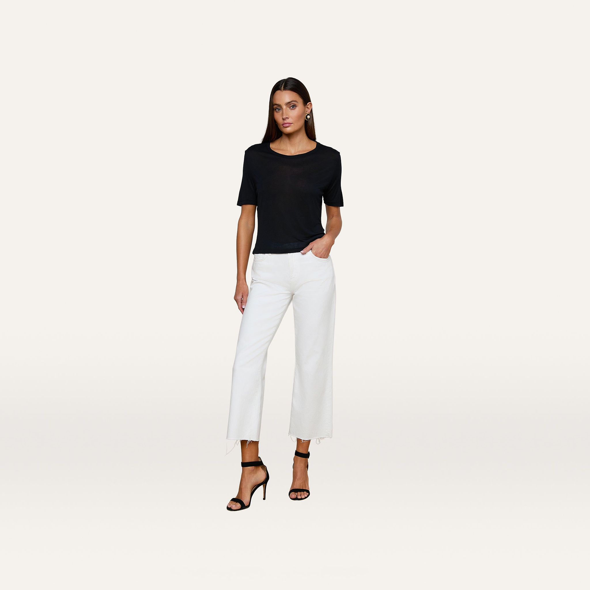 A woman stands against a plain white background, wearing the Remy Tee—a black, short-sleeve top with a relaxed fit—paired with white wide-leg pants and black high-heeled sandals. One hand is in her pocket as she looks directly at the camera.