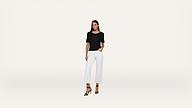 A woman stands against a plain white background, wearing a black short-sleeve top, white cropped pants, and black high-heeled sandals, with one hand in her pocket.