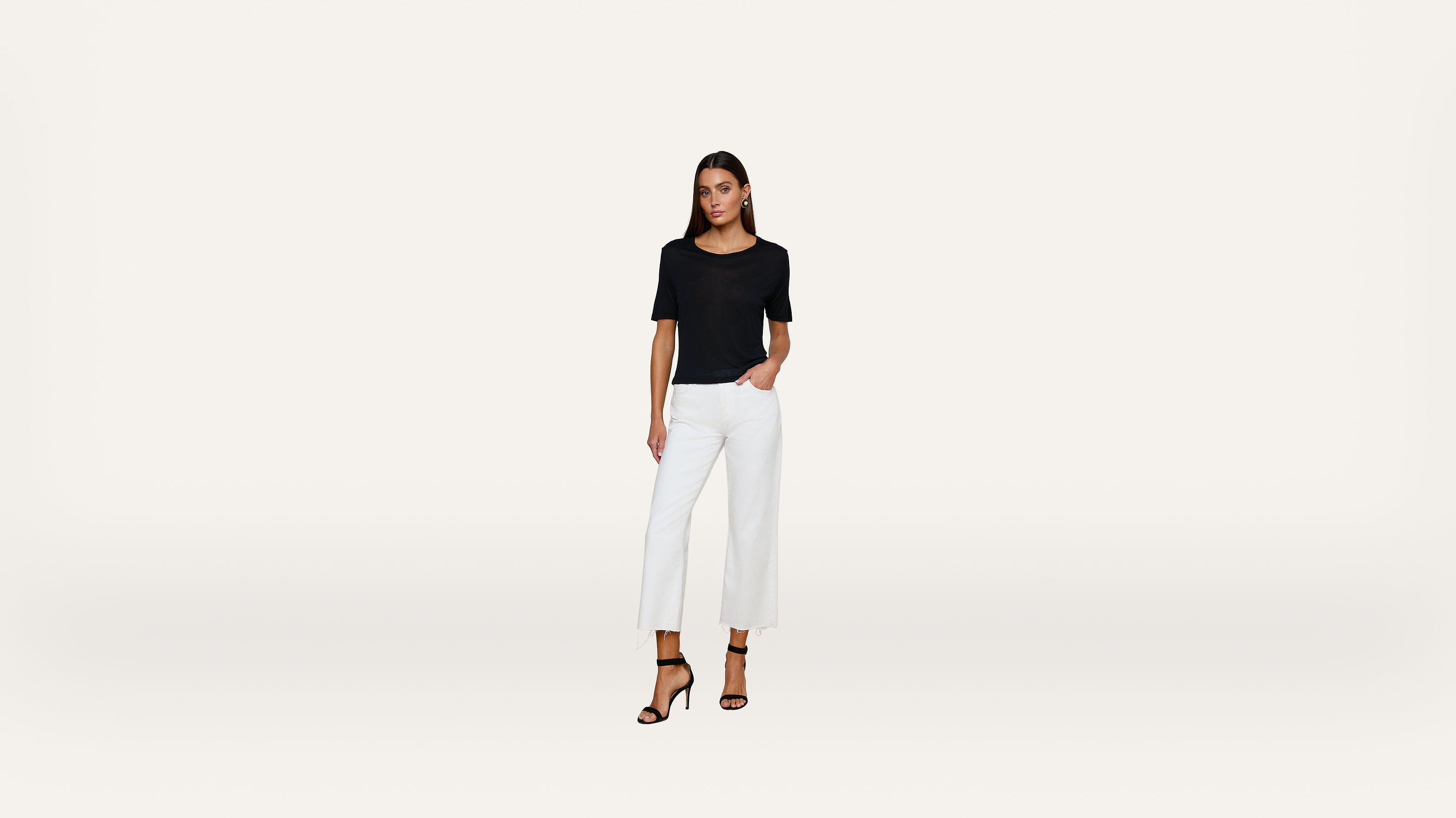 A woman stands against a plain white background, wearing a black short-sleeve top, white cropped pants, and black high-heeled sandals, with one hand in her pocket.
