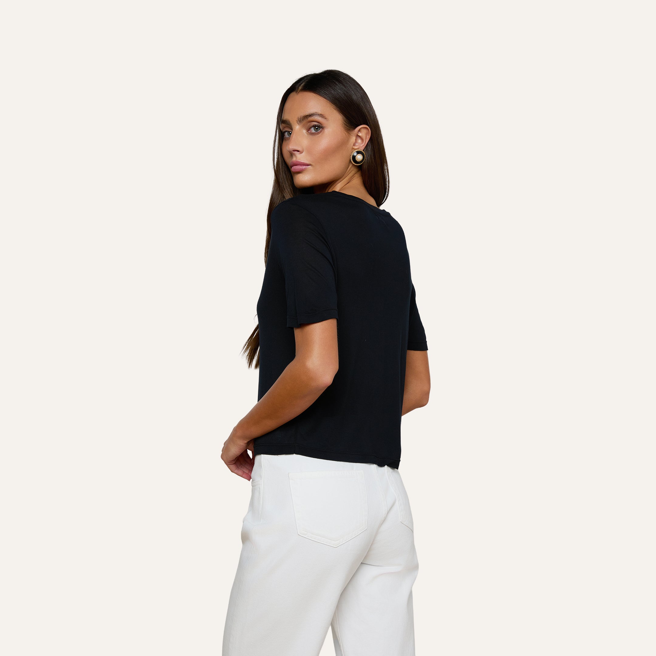 A woman with long straight hair is wearing the Remy Tee, a relaxed fit black short-sleeve top, paired with white pants as she stands turned slightly to the side and looks over her shoulder against a plain light background.