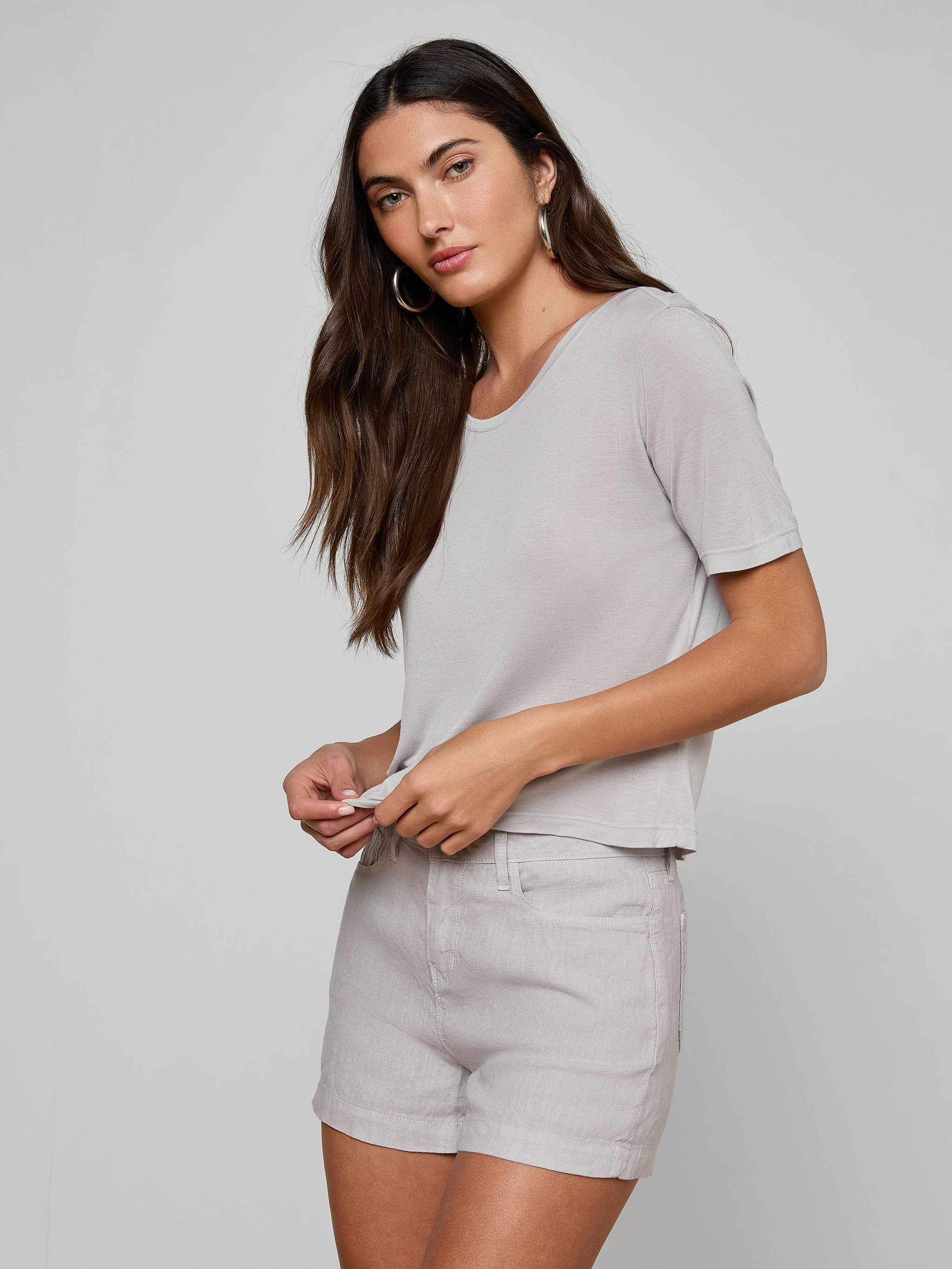 A woman with long brown hair wears the Remy Tee in light gray, paired with matching relaxed fit shorts, standing against an off-white background and gazing slightly to the side with a neutral expression.
