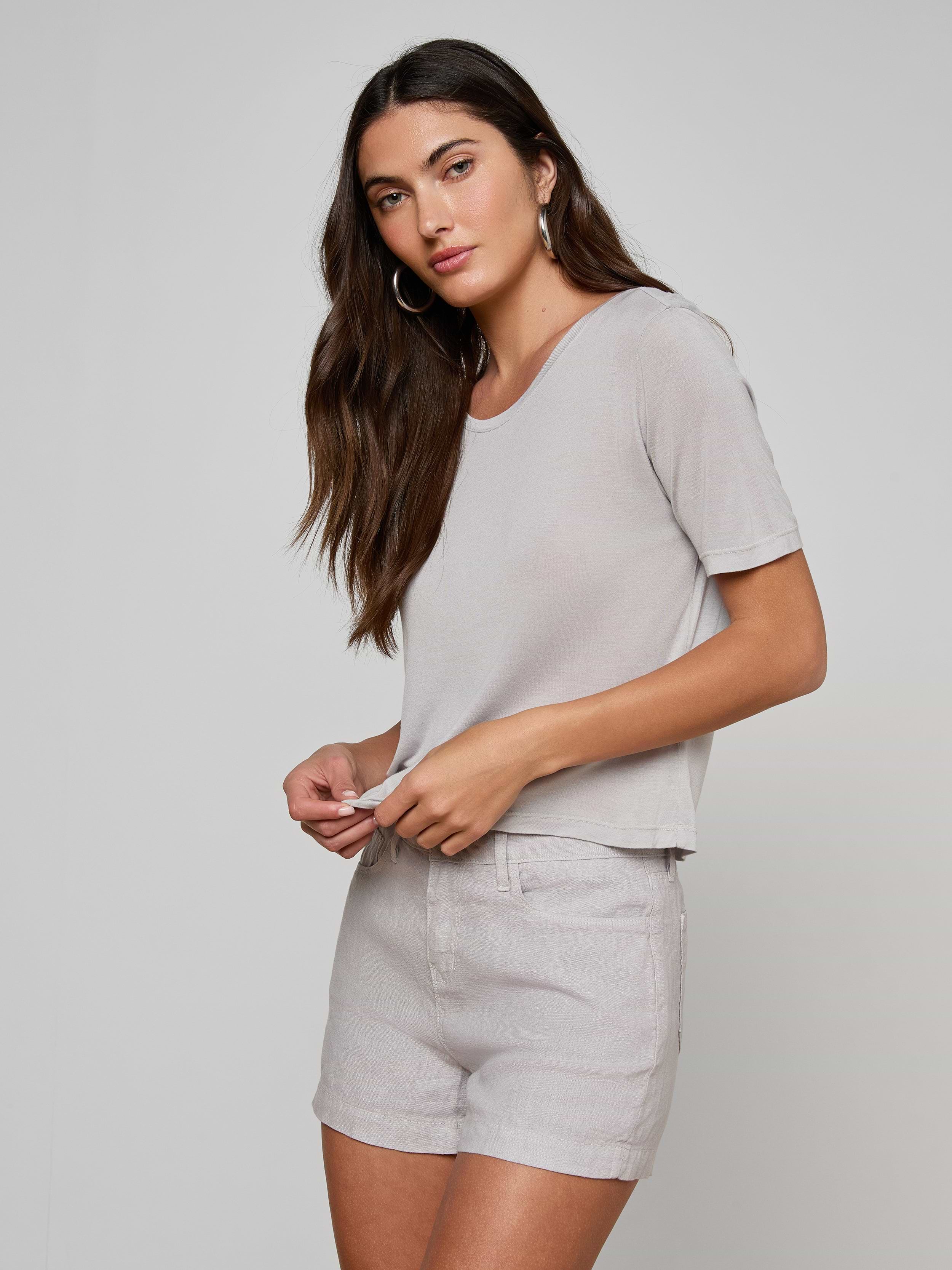 A woman with long brown hair wears the Remy Tee in light gray, paired with matching relaxed fit shorts, standing against an off-white background and gazing slightly to the side with a neutral expression.