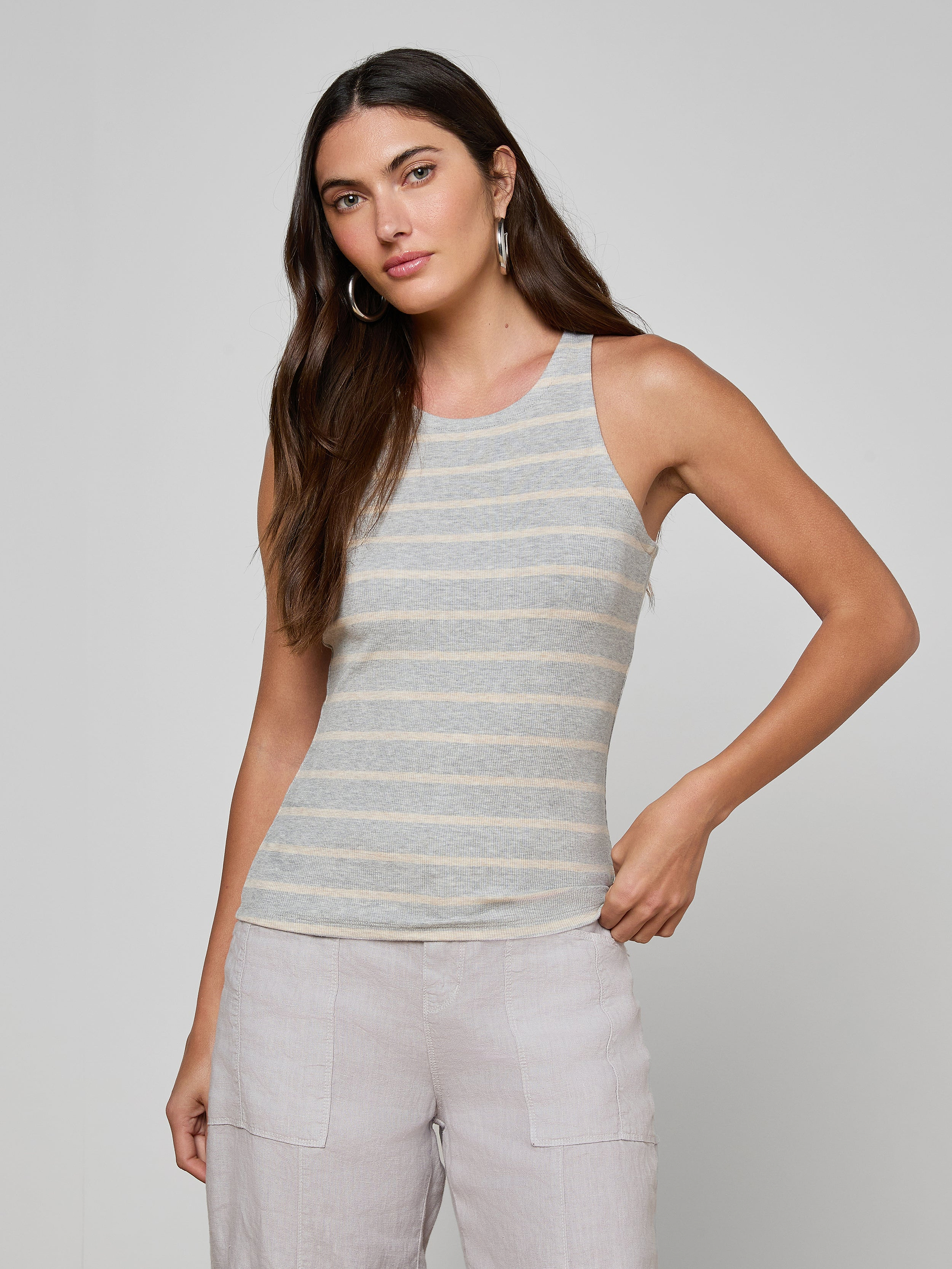 A woman with long brown hair, hoop earrings, and light-colored pants stands against a plain background wearing the Nia Ribbed Tank in gray and white stripes. She faces the camera with a relaxed posture.