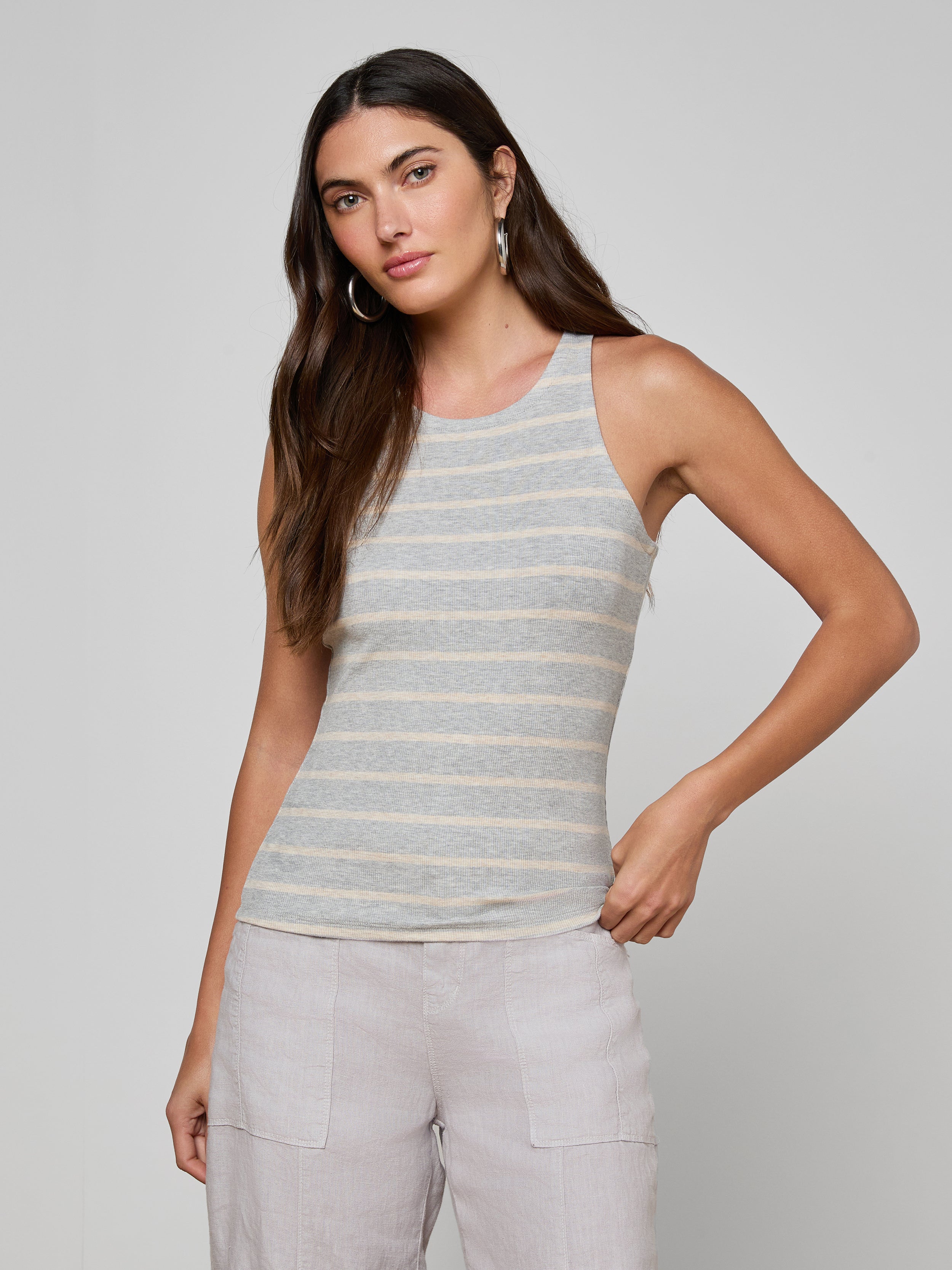 A woman with long brown hair, hoop earrings, and light-colored pants stands against a plain background wearing the Nia Ribbed Tank in gray and white stripes. She faces the camera with a relaxed posture.