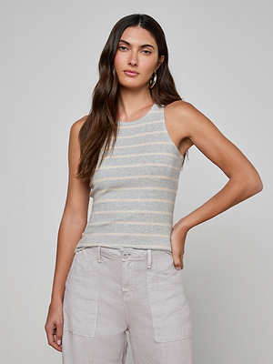 A woman with long brown hair wears the Nia Ribbed Tank, a sleeveless gray and white striped microfiber modal top, paired with high-waisted light pants. She stands against a plain background with one hand on her hip.