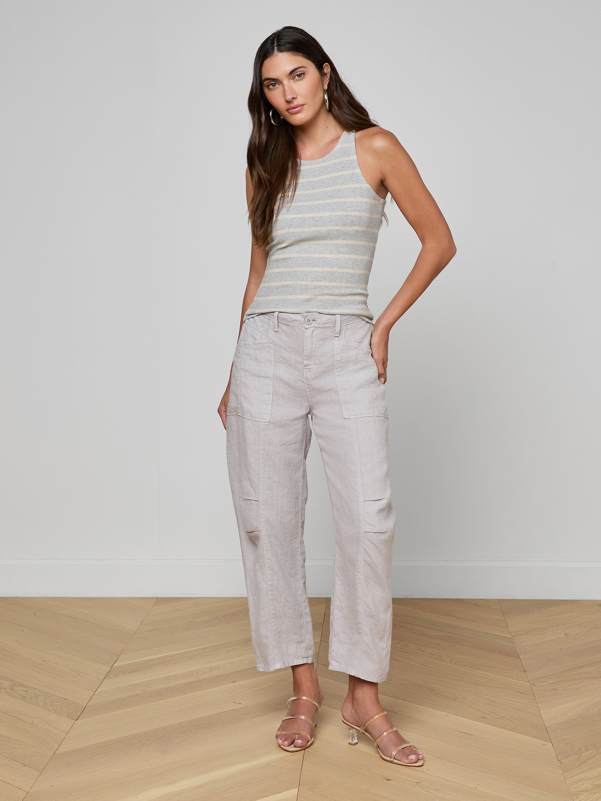 A woman with long hair down poses indoors on a wooden floor, wearing the Nia Ribbed Tank, light wide-leg pants with pockets, and strappy sandals. She stands with one hand in her pocket.
