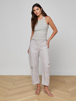 A woman with long hair down poses indoors on a wooden floor, wearing the Nia Ribbed Tank, light wide-leg pants with pockets, and strappy sandals. She stands with one hand in her pocket.