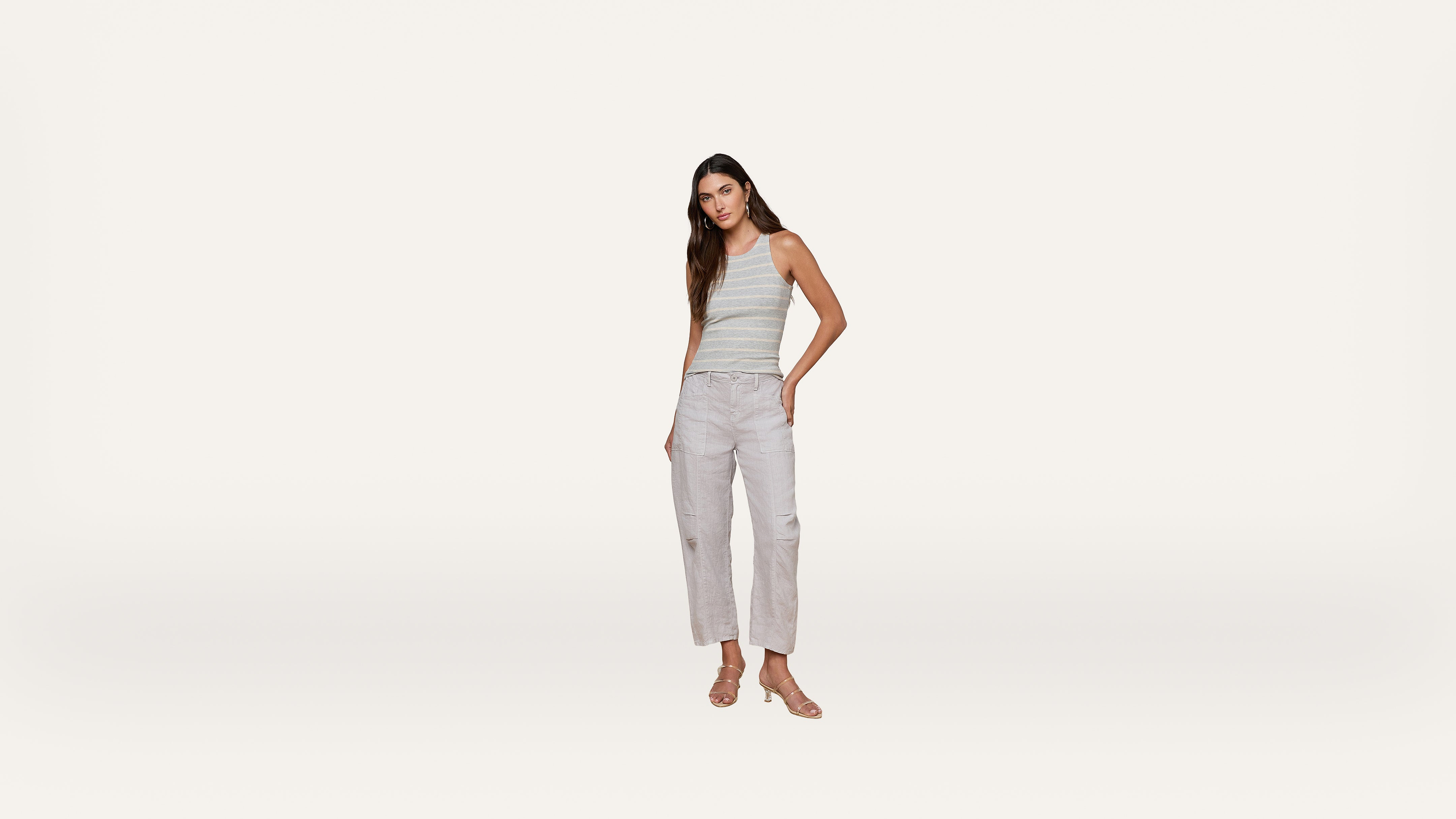 A woman stands against a plain white background, wearing a striped sleeveless top, light gray cropped pants, and sandals. One hand is in her pocket, and she looks toward the camera.