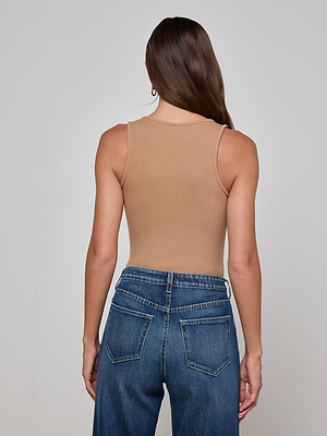 A woman with brown hair stands facing away in a fitted beige Adira Tank by L'AGENCE and high-waisted blue jeans. Her hair is parted to one side and falls down her back. The background is plain and light-colored.