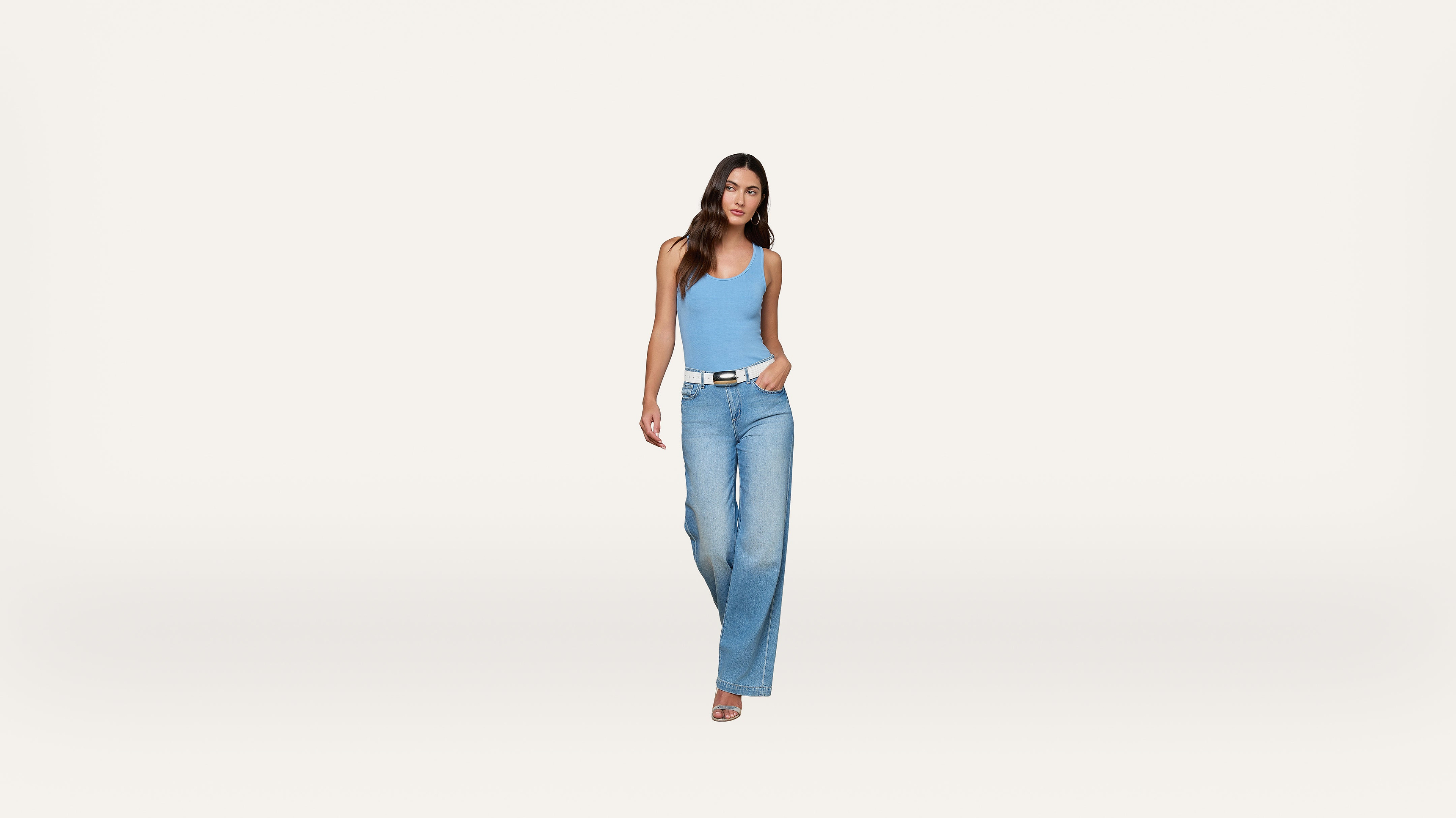 A woman stands against a plain white background, wearing a light blue tank top, wide-leg blue jeans, a white belt, and sandals. One hand is in her pocket and she is looking forward with a neutral expression.