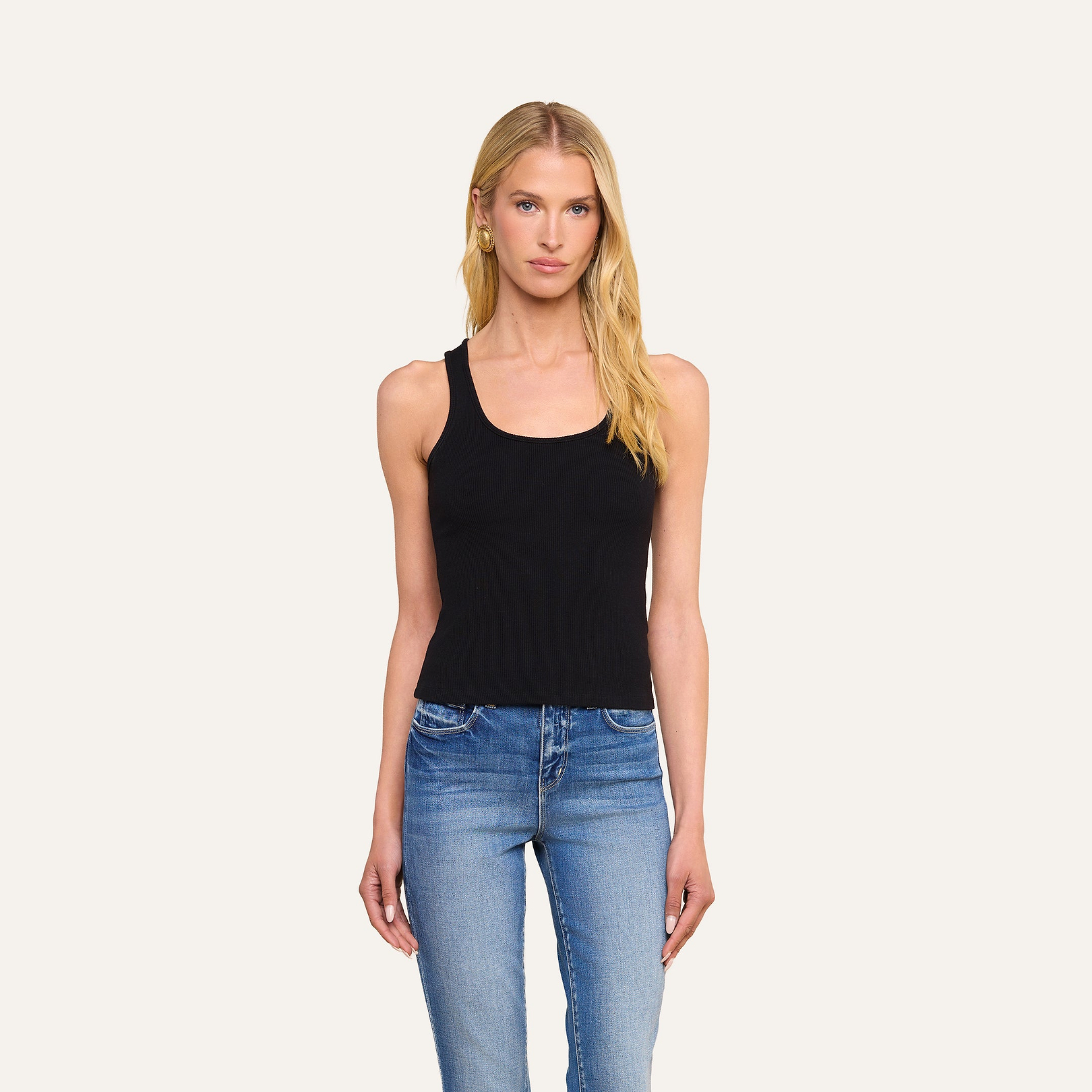 Una mujer con el pelo largo y rubio posa frente a un fondo liso de color beige, mirando a la cámara con una expresión neutra, vestida con la camiseta sin mangas Adira Scoopneck en color negro y unos vaqueros azules.