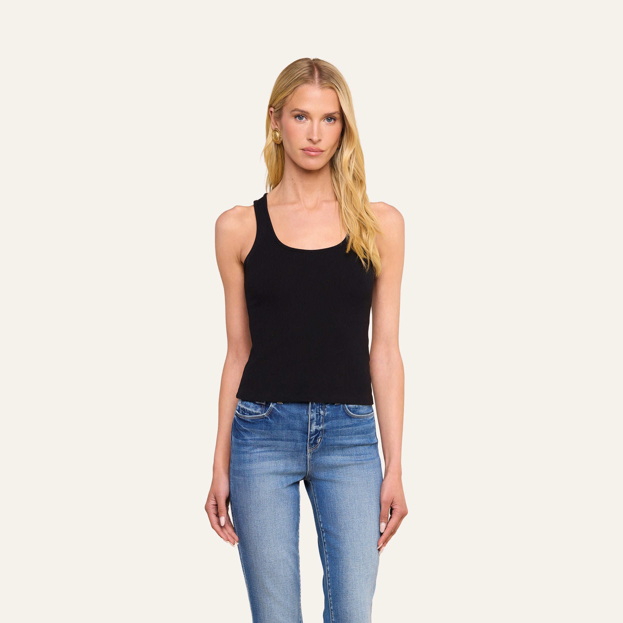 A woman with long blonde hair wears the Adira Scoopneck Tank in black with blue jeans, standing against a plain light background, facing forward with a neutral expression.