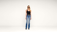 A woman wearing sunglasses, a black tank top, blue jeans, and black heels stands against a plain white background.
