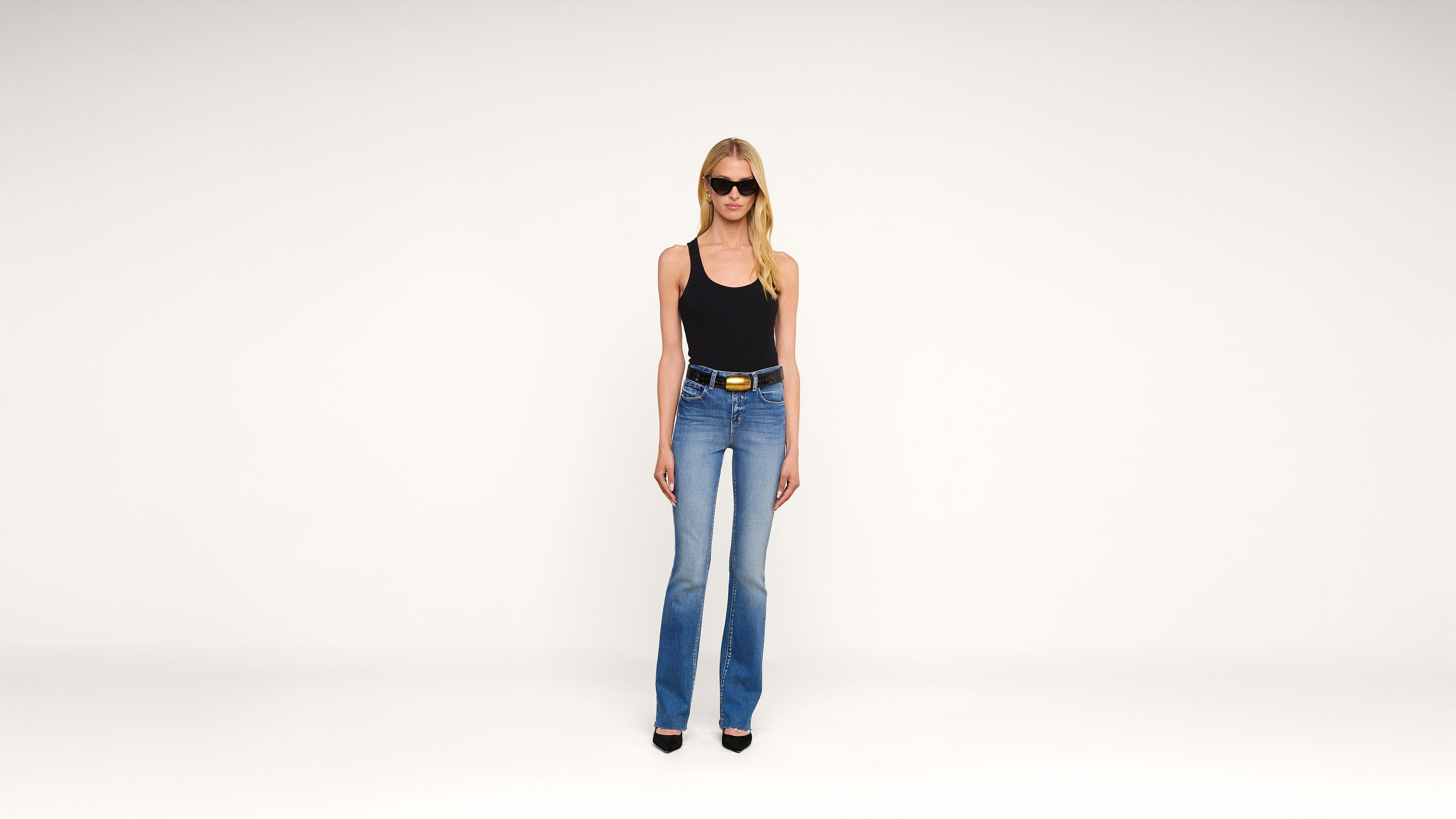 A woman wearing sunglasses, a black tank top, blue jeans, and black heels stands against a plain white background.