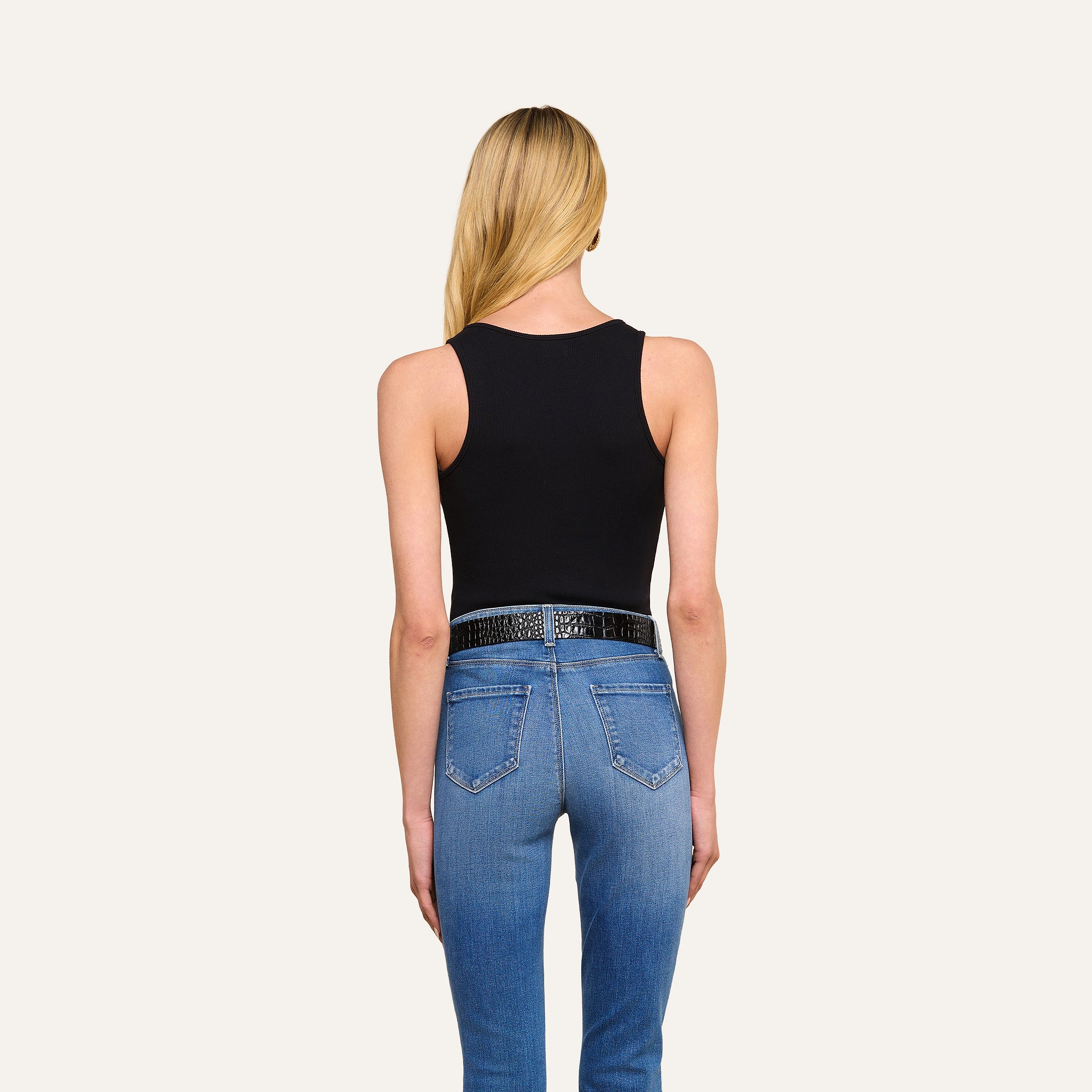 A woman with long blonde hair, dressed in the Adira Scoopneck Tank and blue slim fit jeans with a black belt, stands facing away from the camera against a plain, light background.