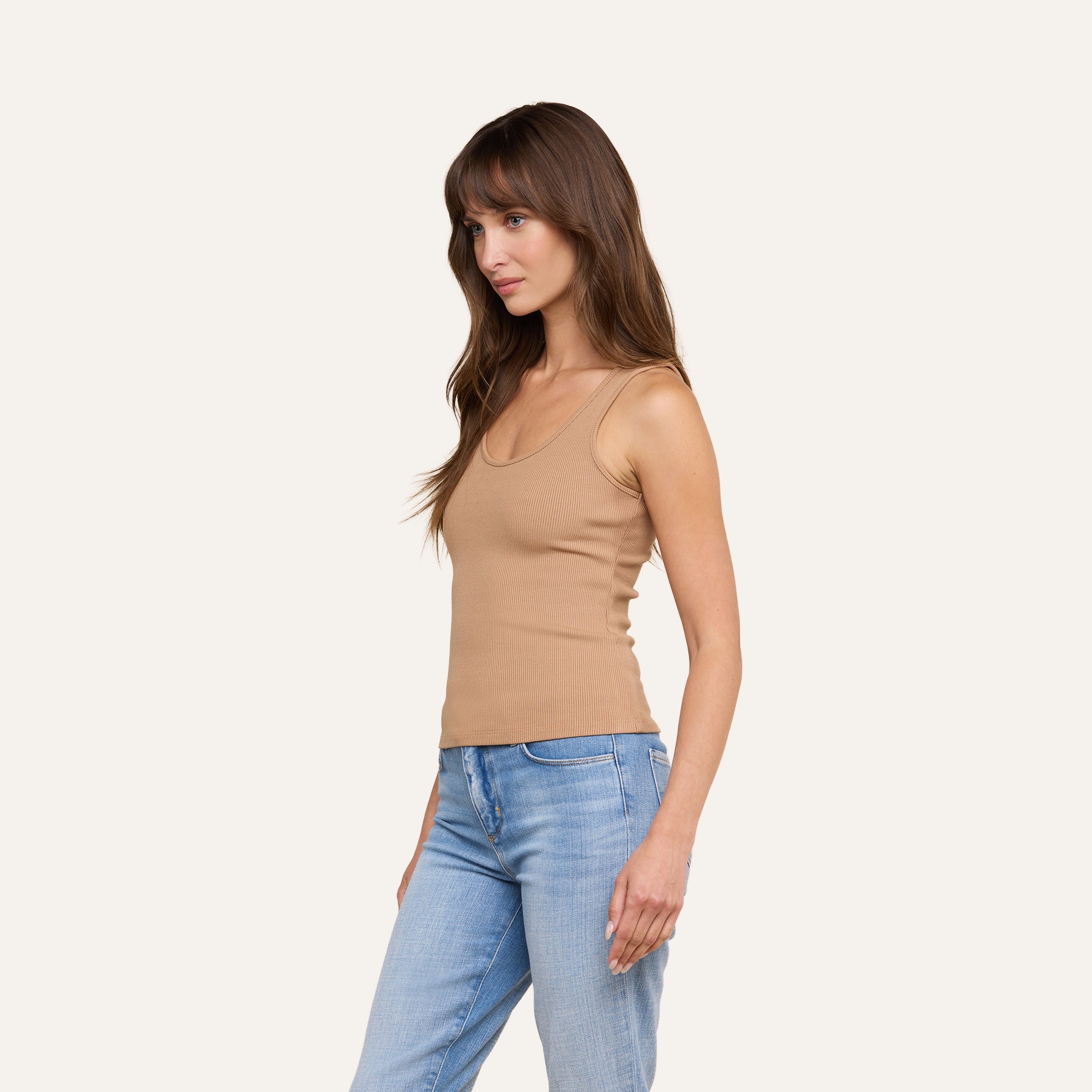 Eine Frau mit langen braunen Haaren steht seitlich im Bild und trägt das Adira-Tanktop mit tiefem U-Ausschnitt sowie hellblaue Jeans vor einem schlichten beigen Hintergrund.