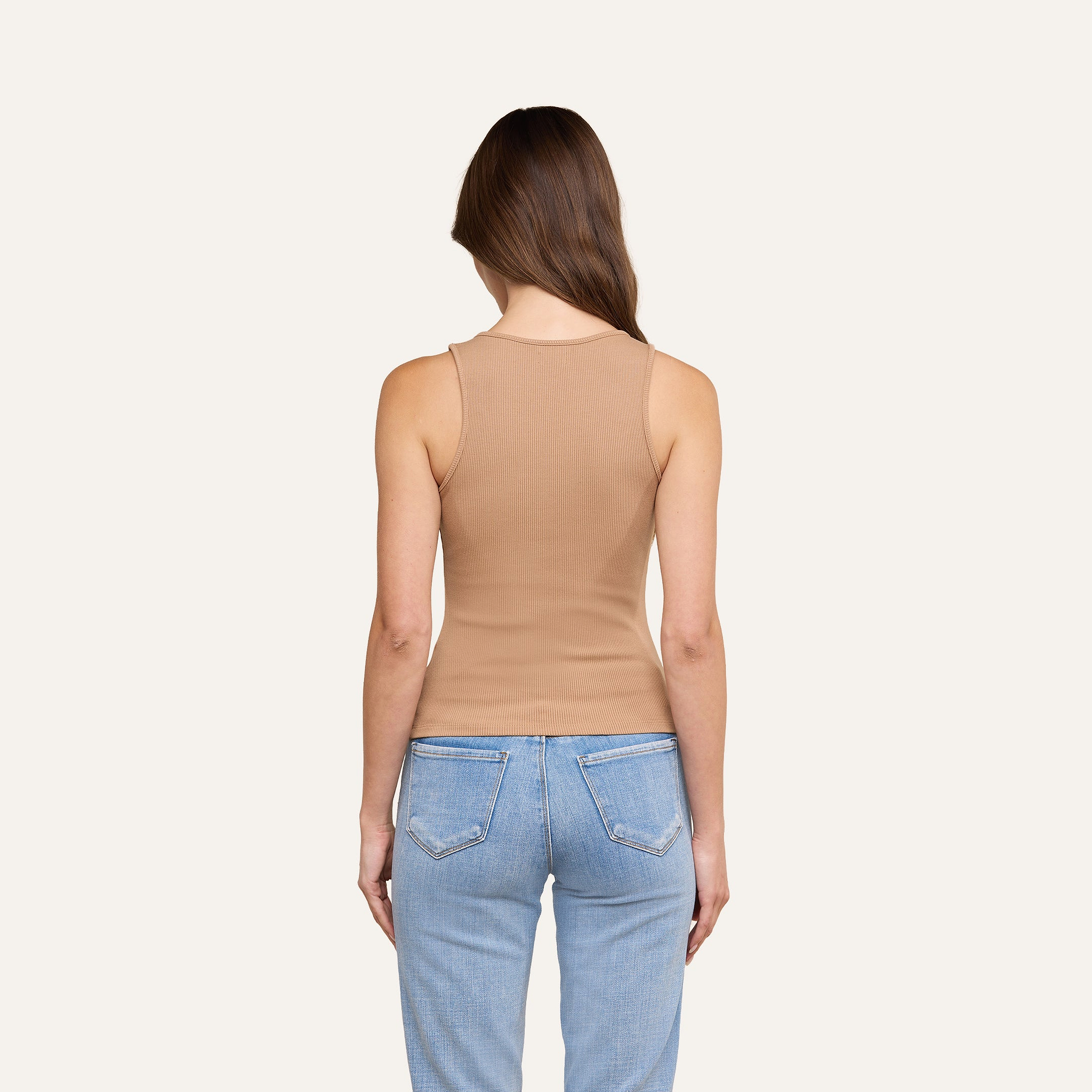 Eine Frau mit langen braunen Haaren steht mit dem Rücken zur Kamera und trägt das eng anliegende, gerippte Adira-Tanktop sowie hellblaue Jeans vor einem schlichten beigen Hintergrund.