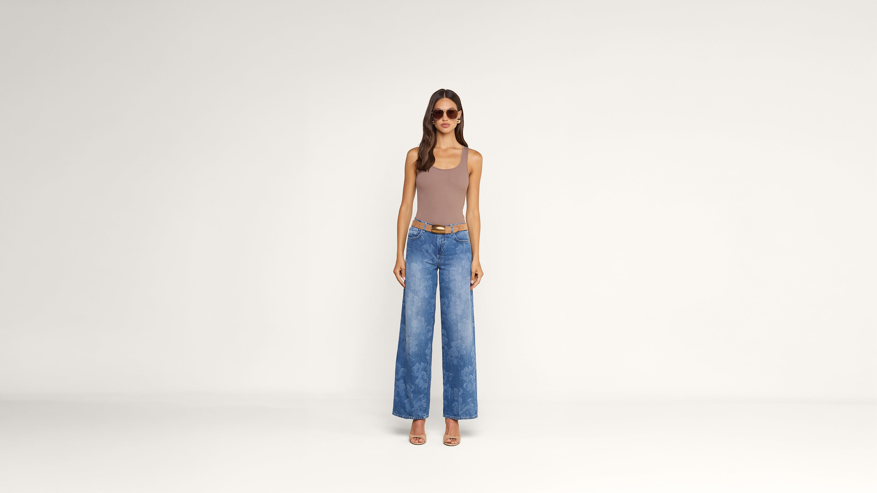 A woman wearing sunglasses, a brown sleeveless top, blue high-waisted wide-leg jeans, and sandals stands against a plain white background.