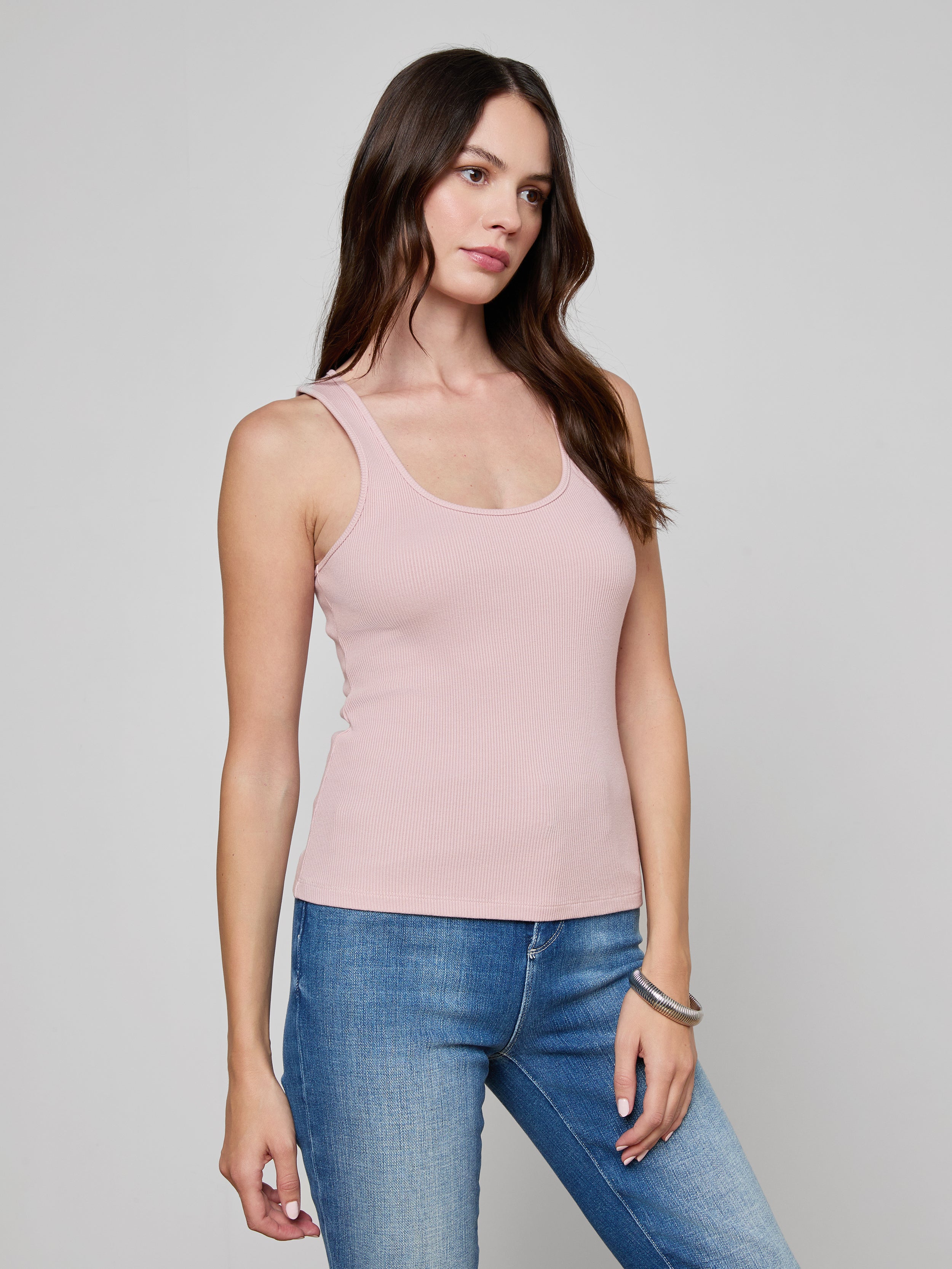 A woman with long brown hair wears the Adira Scoopneck Tank in light pink and blue jeans, standing against a plain light background with a neutral expression.