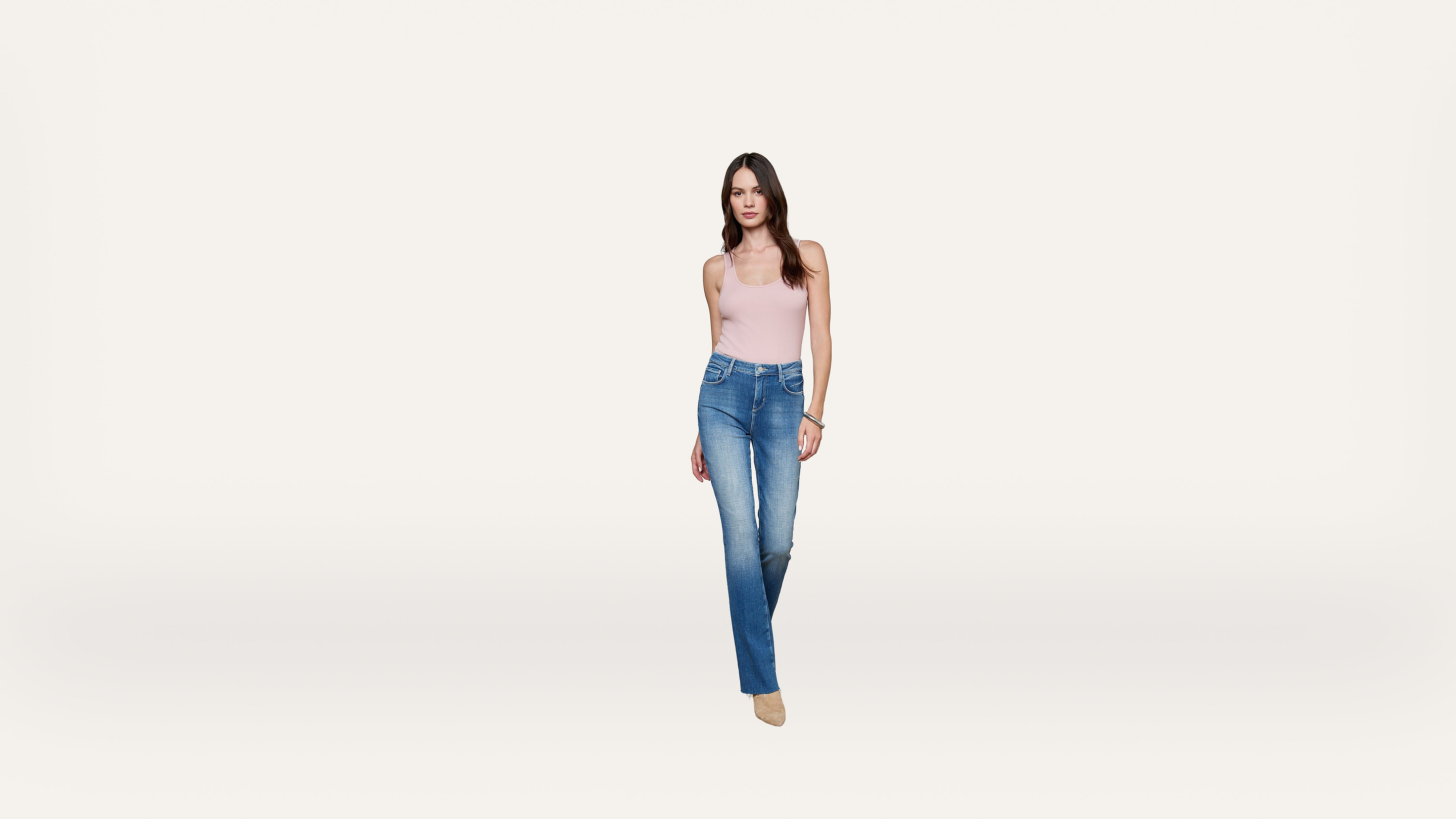 A woman wearing a light pink tank top, blue jeans, and beige shoes stands against a plain white background, facing forward.