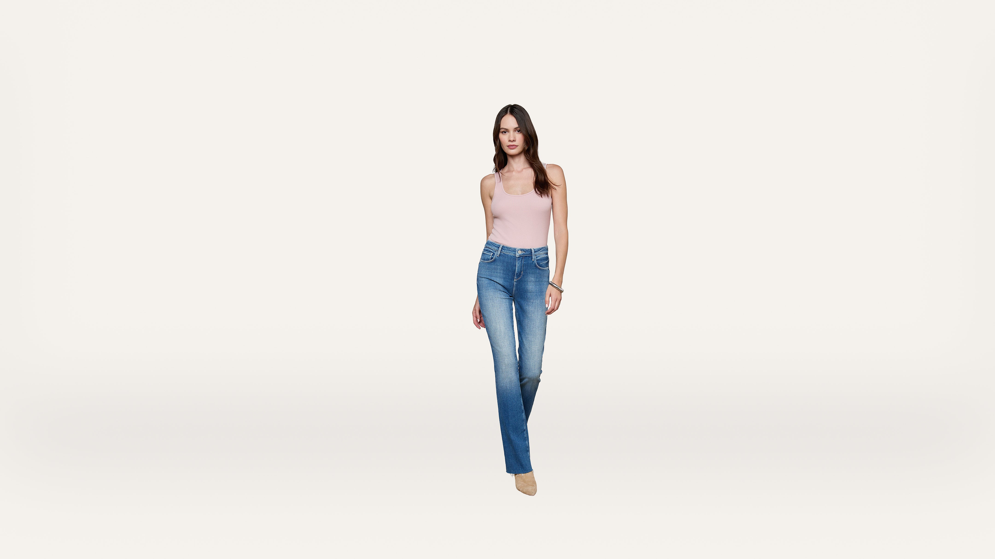 A woman wearing a light pink tank top, blue jeans, and beige shoes stands against a plain white background, facing forward.