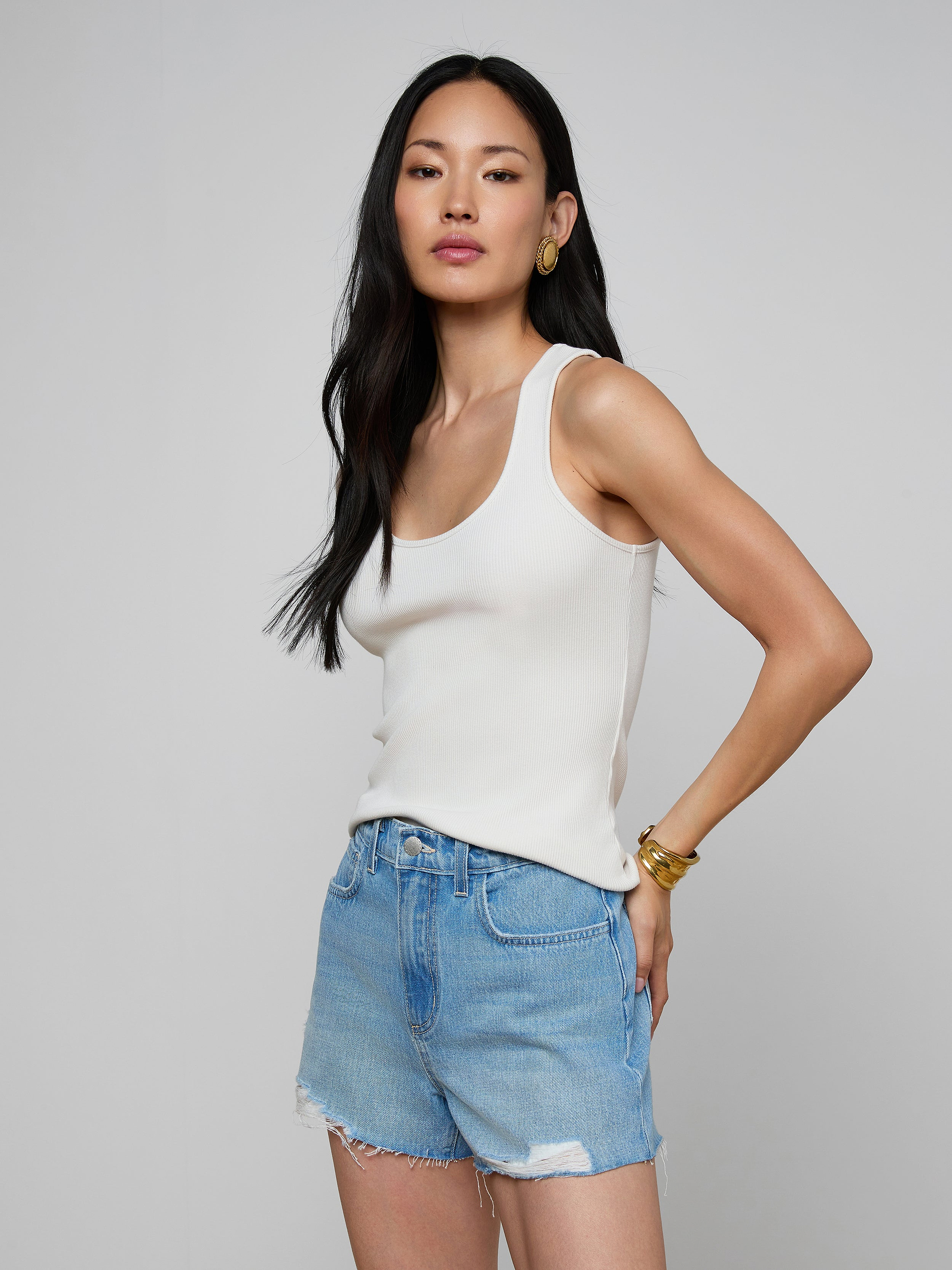 A woman stands against a plain background, wearing the Adira Scoopneck Tank in white, beige pants with a belt, and gold earrings. She has long blonde hair and a neutral expression, facing slightly to the left.