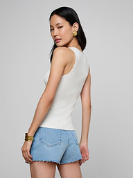 A woman with long blonde hair, wearing the Adira Scoopneck Tank in white and light beige pants, stands facing away from the camera against a plain light background.