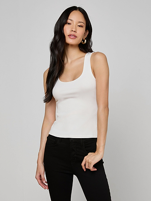 A woman with long black hair wears the Adira Scoopneck Tank in white and black pants, standing against a plain light gray background and looking confidently at the camera with a relaxed posture.