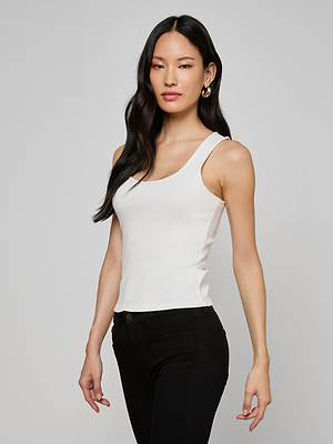 A woman with long black hair wears the slim fit ribbed knit Adira Scoopneck Tank in white and black pants, standing against a plain light gray background and looking confidently at the camera.