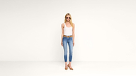 A woman wearing sunglasses, a white tank top, blue jeans, brown belt, and brown shoes stands against a plain white background.