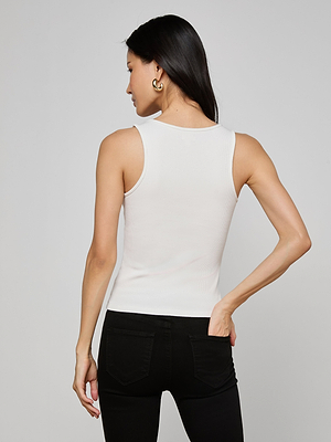 A woman with long dark hair is seen from the back, wearing the Adira Scoopneck Tank in white with black jeans and gold hoop earrings, her hand on her hip against a plain light background.