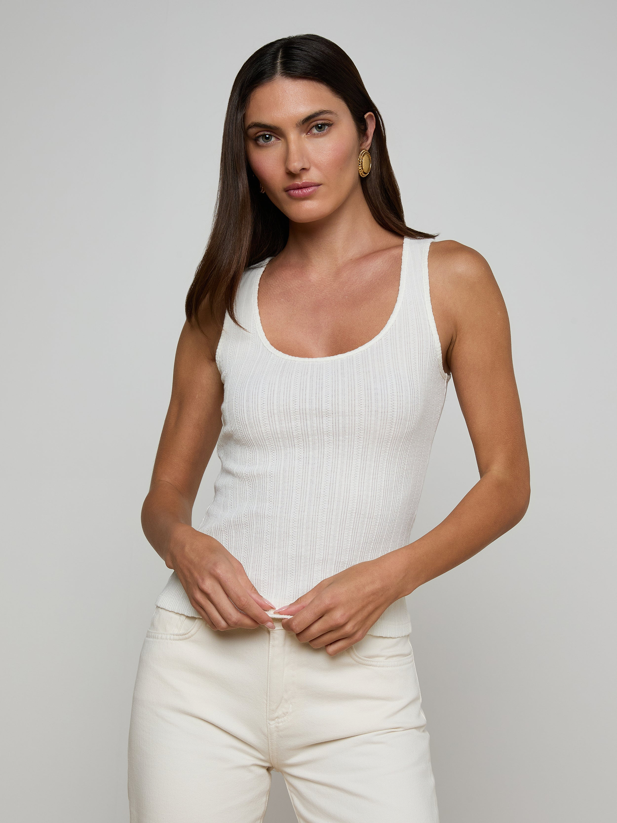 Woman with long brown hair wearing the Adira Scoopneck Tank and off-white pants, standing against a plain light background, looking at the camera with a neutral expression.