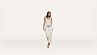 A woman with long dark hair wearing a white tank top, wide-leg white pants, a brown belt, and beige heels walking forward against a plain white background.