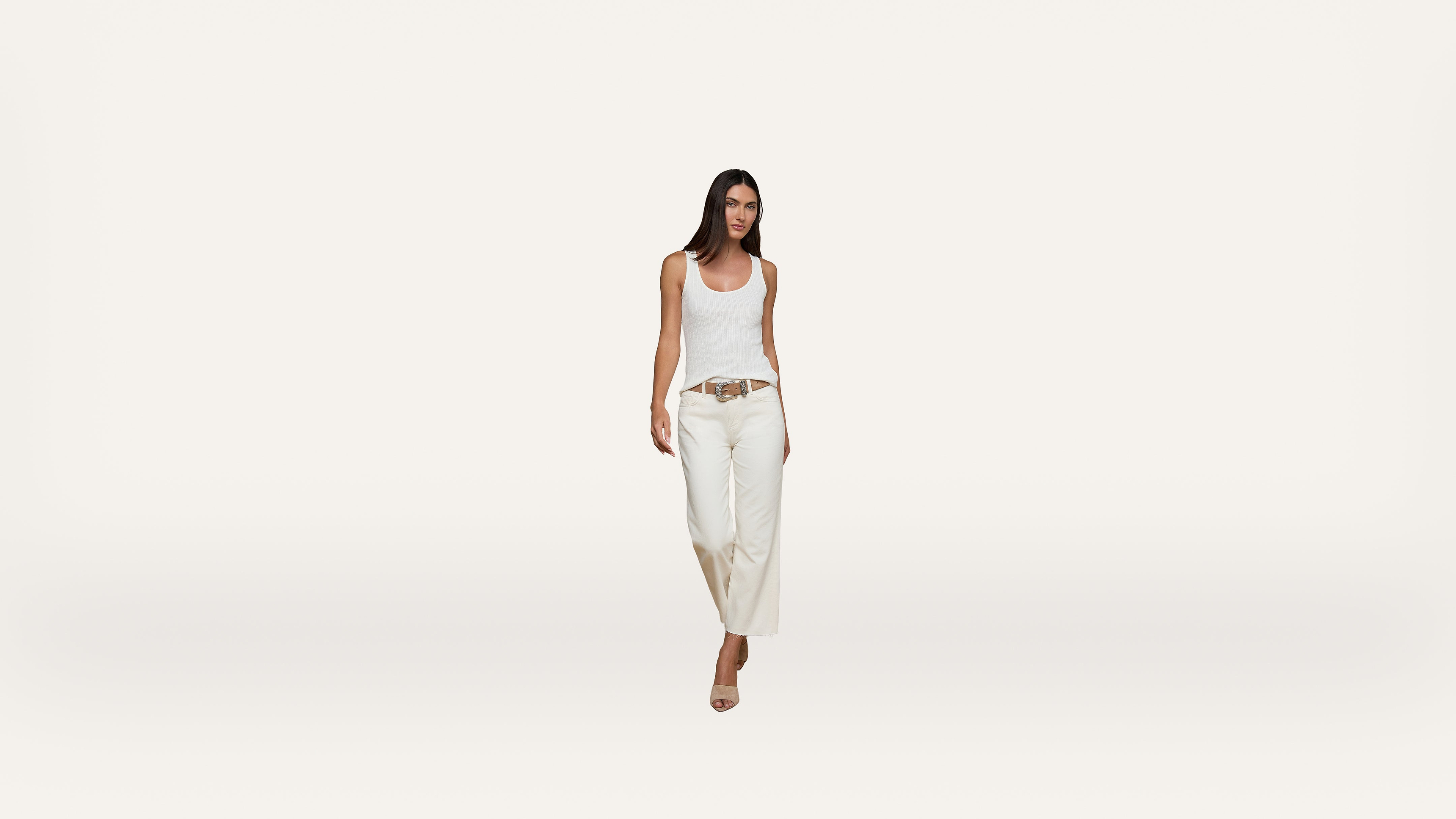 A woman with long dark hair wearing a white tank top, wide-leg white pants, a brown belt, and beige heels walking forward against a plain white background.