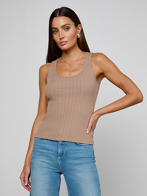 A woman with long brown hair wears the Adira Scoopneck Tank in beige, paired with blue jeans and gold jewelry, standing against a plain light gray background with a neutral expression.