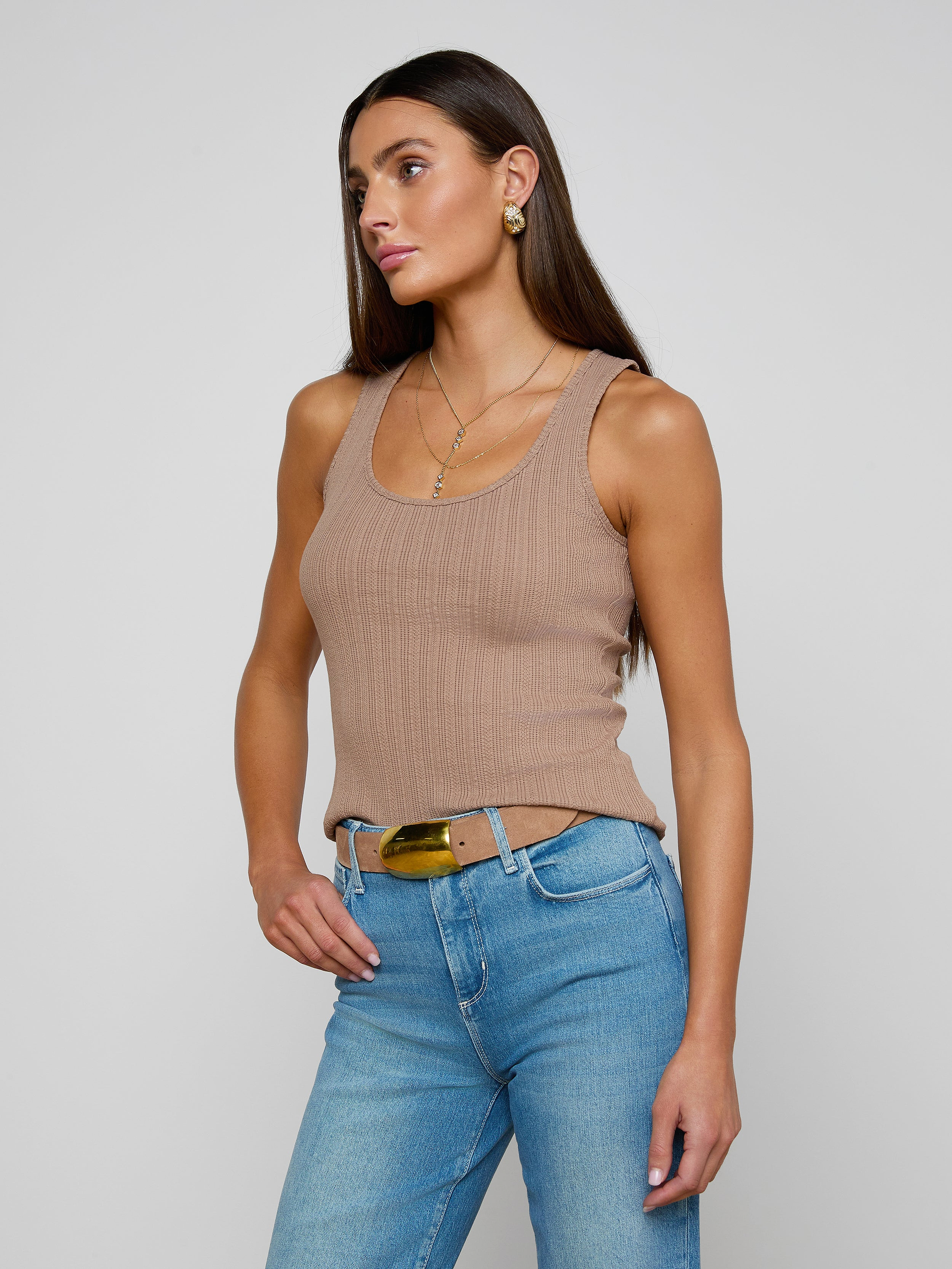 Wearing the Adira Scoopneck Tank—a fitted tan ribbed top—paired with blue jeans and a belt, a woman stands against a plain light background, gazing slightly to the side with one hand in her pocket.