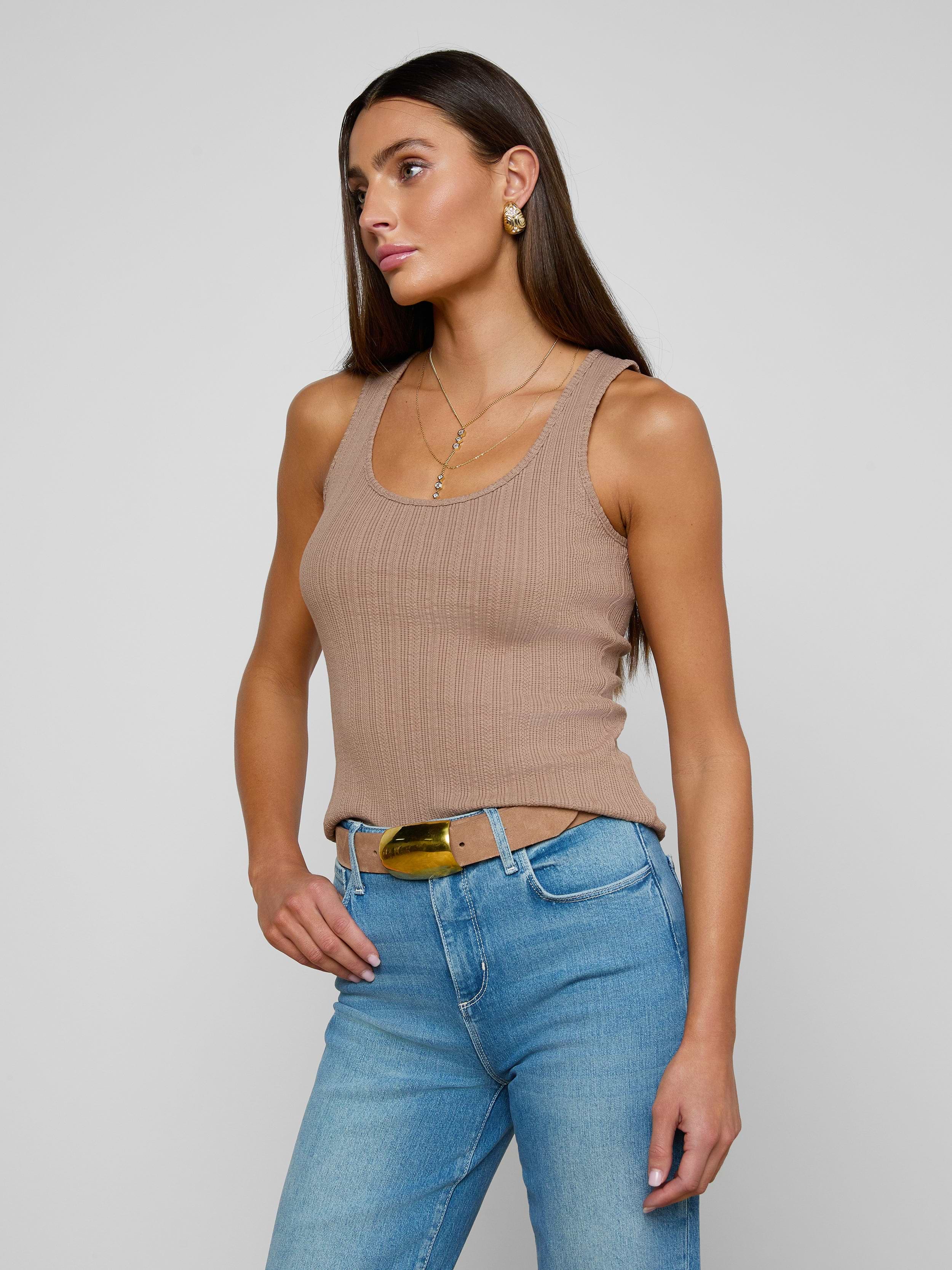 Wearing the Adira Scoopneck Tank—a fitted tan ribbed top—paired with blue jeans and a belt, a woman stands against a plain light background, gazing slightly to the side with one hand in her pocket.