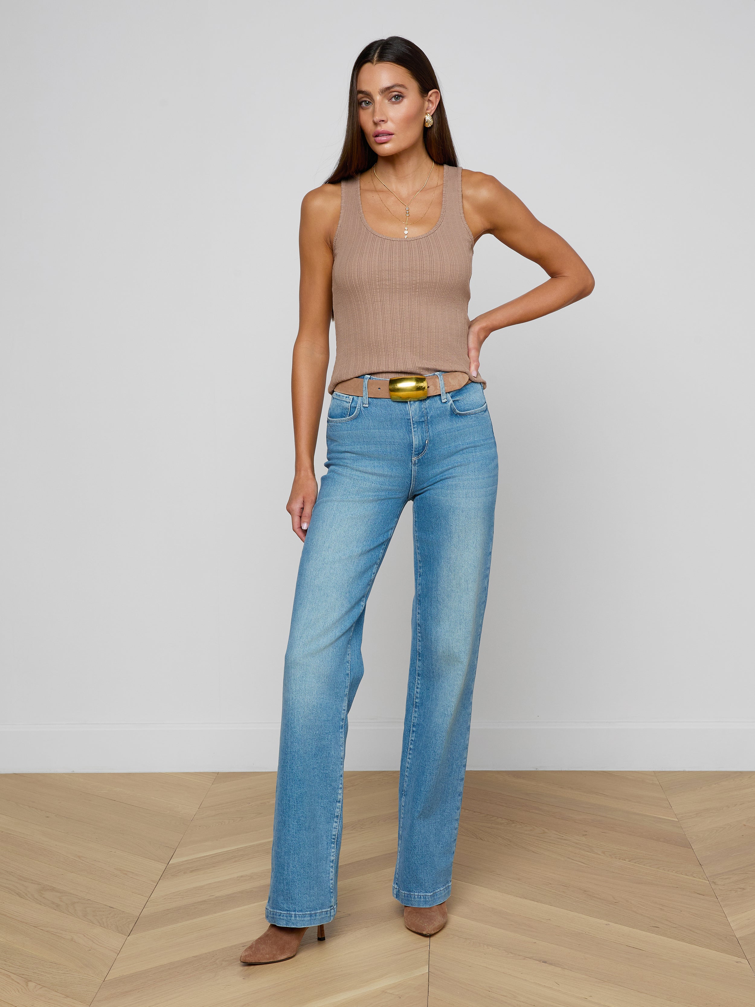A woman models the Adira Scoopneck Tank— a fitted, sleeveless beige top with a scoop neckline— paired with blue jeans and brown heels, standing with one hand on her hip against a plain white background.