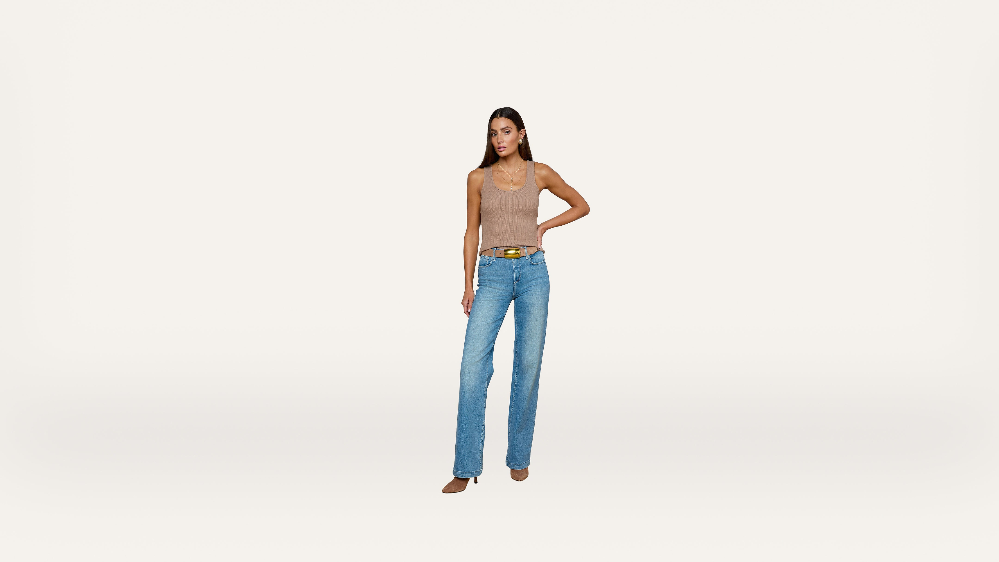 A woman in a beige sleeveless top and light blue jeans stands with one hand on her hip against a plain white background.