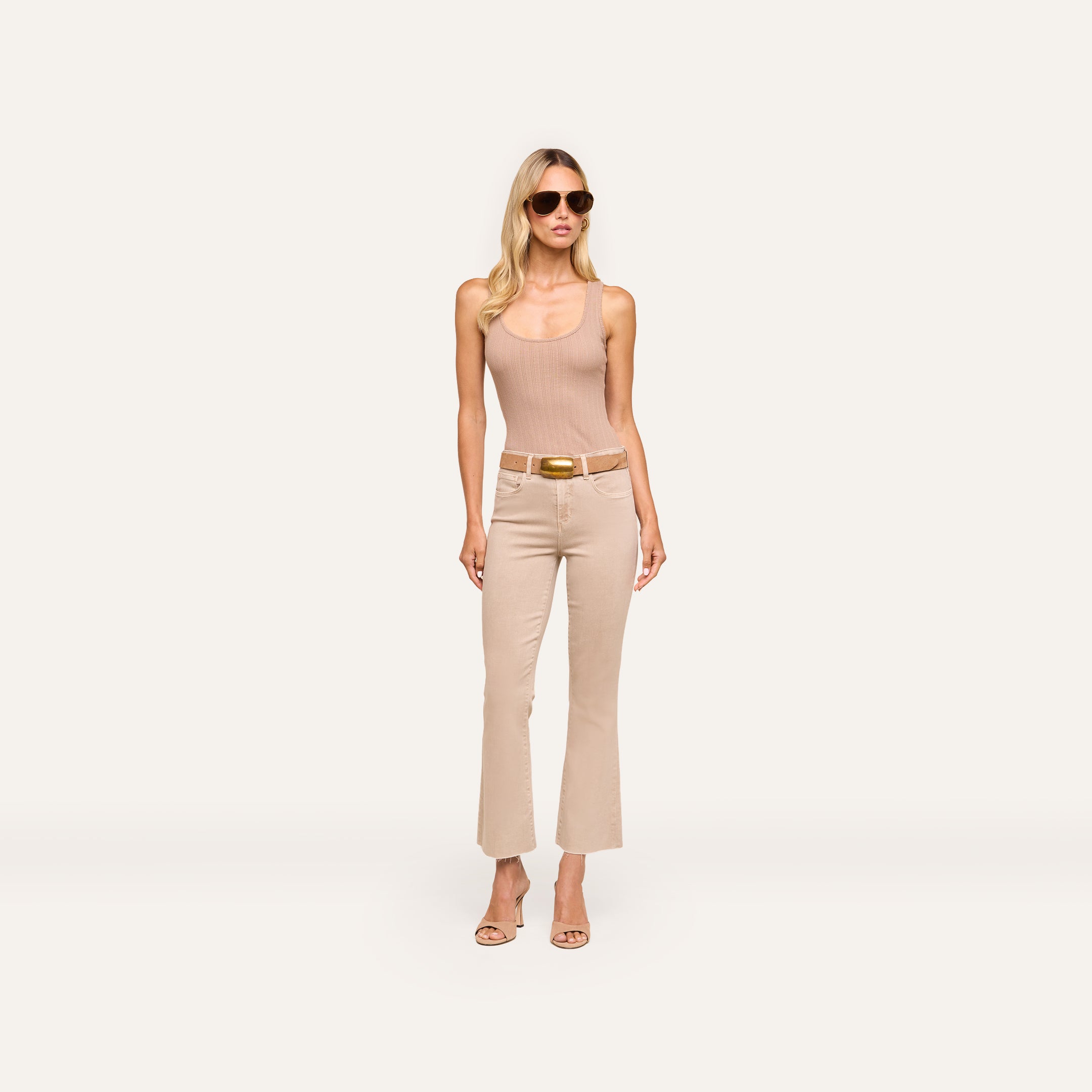 Woman wearing sunglasses, the Adira Scoopneck Tank in organic cotton, beige belted pants, and heeled sandals stands against a plain light background, looking forward with hands by her sides.