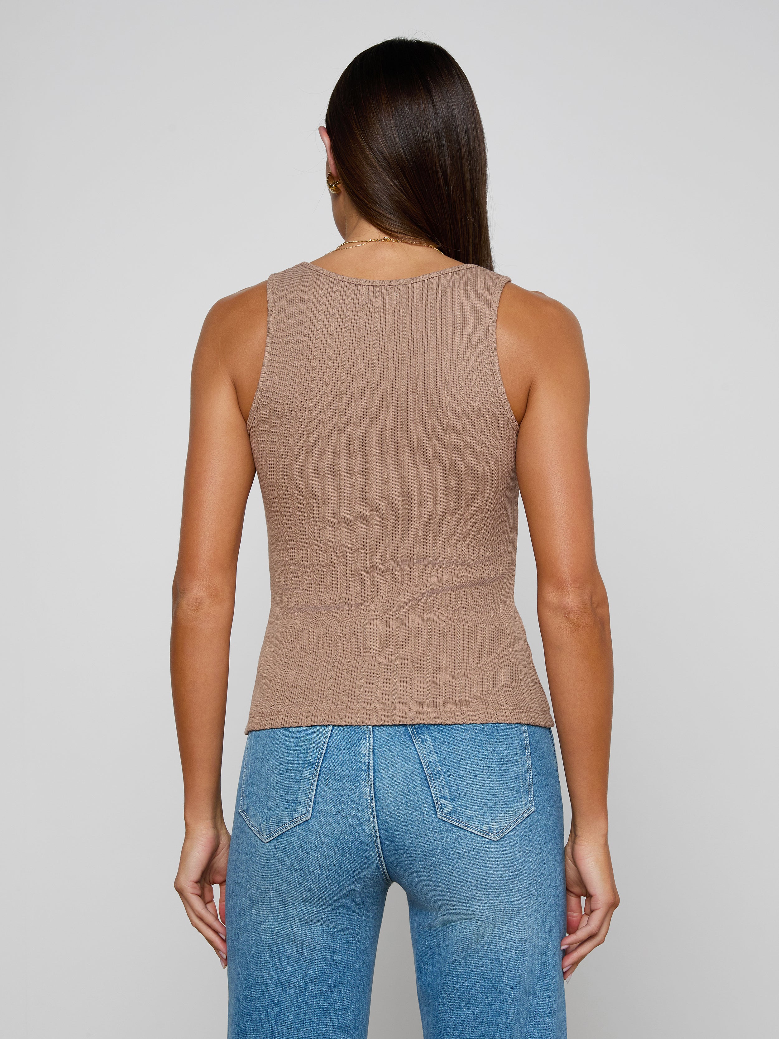 A woman with long brown hair, wearing the Adira Scoopneck Tank—a fitted, sleeveless beige top made from organic cotton—and blue jeans stands with her back to the camera against a plain light background.