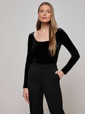 A woman with long, straight, light brown hair wears the L'AGENCE Jelena Velvet Top—slim fit, black stretch velvet with a wide U-neck—paired with black pants. She stands against a plain light gray background, one hand in her pocket.