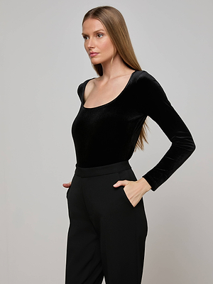 A woman with long, straight brown hair wears the L'AGENCE Jelena Velvet Top in black, paired with high-waisted black pants, as she stands with her hands in her pockets against a plain light background.